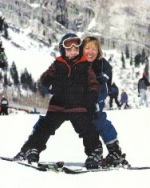 Adult behind child doing adaptive skiing with a snowy mountain background